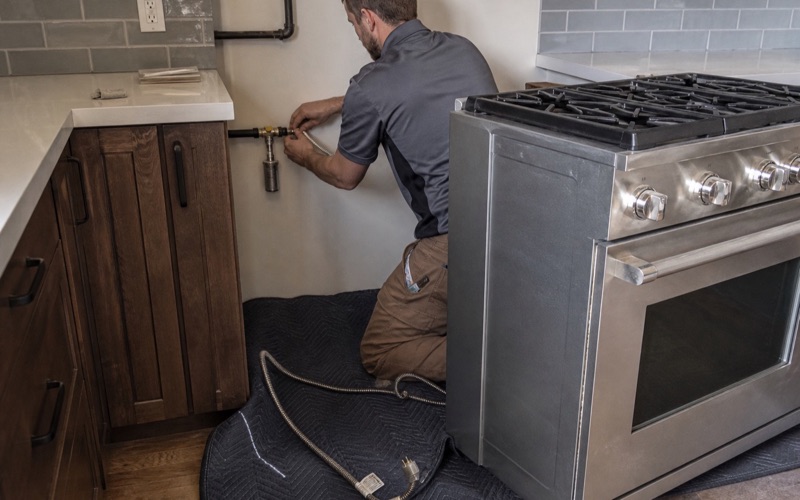 An installer connecting a flexible gas connector behind a pulled-out kitchen range
